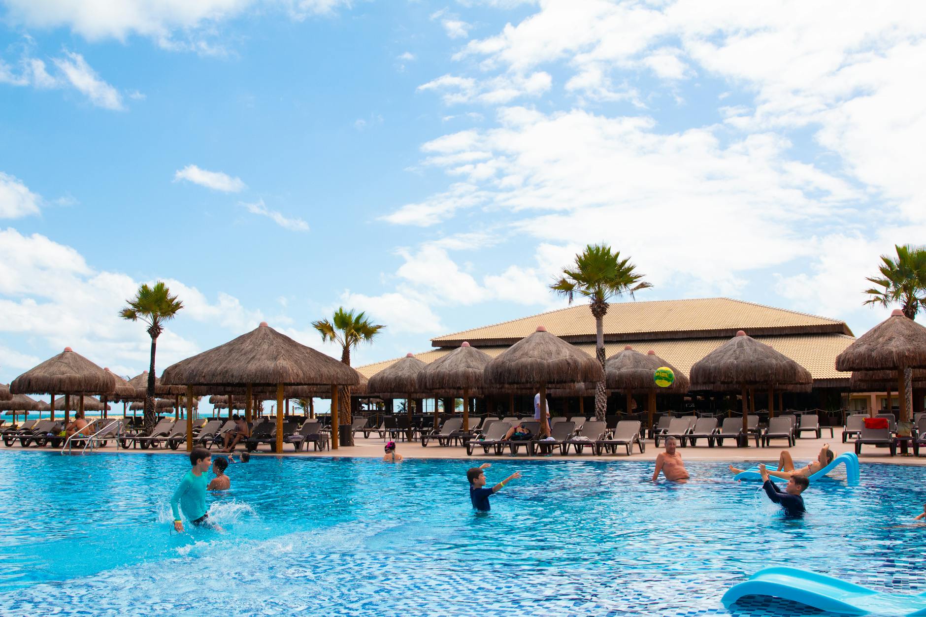 Families enjoying a sunny day at a resort pool with palm trees and thatched umbrellas